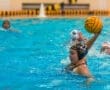 Water polo player wearing a black cap with number 22, preparing to shoot the yellow ball amidst splashing water.