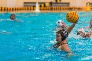 Water polo player wearing a black cap with number 22, preparing to shoot the yellow ball amidst splashing water.