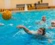 Player reaching for a water polo ball during a match, with teammates and splashes visible in the background.