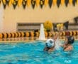 Water polo players compete in a pool, one aiming to throw a ball while the other defends. Colorful banners decorate the scene.