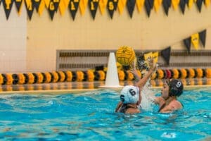 Water polo players compete in a pool, one aiming to throw a ball while the other defends. Colorful banners decorate the scene.