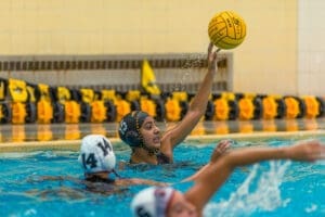 A water polo player in a black cap throws a ball amidst teammates in a swimming pool. Sprays of water surround them.