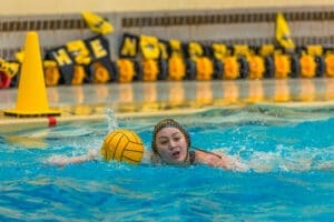 Water polo athlete focuses on the game while swimming, with a yellow ball in play and an intense competitive atmosphere.