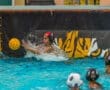 Water polo player deflects a ball at the goal during a match, with teammates observing in the pool's vibrant water.