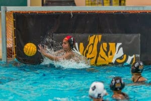 Water polo player deflects a ball at the goal during a match, with teammates observing in the pool's vibrant water.