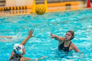 Water polo players compete in a fast-paced game, one athlete passing the ball while another reaches to defend.
