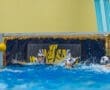 Water polo players competing; one attempts a goal while the goalkeeper dives to block the shot in a dynamic match.