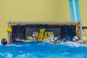 Water polo players competing; one attempts a goal while the goalkeeper dives to block the shot in a dynamic match.