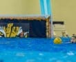 Water polo player focuses on passing a yellow ball, with a goal in the background featuring a tiger design.