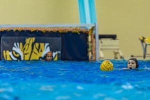 Water polo player focuses on passing a yellow ball, with a goal in the background featuring a tiger design.