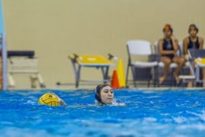Water polo player swims and navigates a game in a pool, with teammates watching from the sidelines.