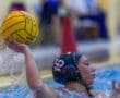 Female water polo player prepares to throw a yellow ball, splashing water in a competitive game setting.