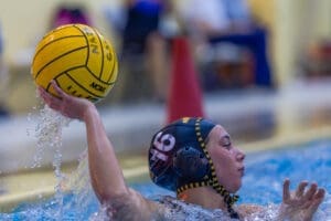 Female water polo player prepares to throw a yellow ball, splashing water in a competitive game setting.