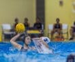 Female water polo player prepares to pass the ball while another player attempts to block her in an indoor pool setting.