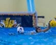 Water polo match action, featuring a player reaching for the ball while another swims nearby, splashing in the water.