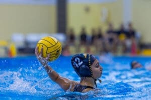 Female water polo player prepares to throw a yellow ball in a pool, showcasing athleticism amidst splashing water.
