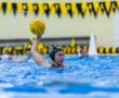 A female water polo player prepares to throw a game ball in a competitive match, with splashes and vibrant surroundings.