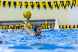 A female water polo player prepares to throw a game ball in a competitive match, with splashes and vibrant surroundings.