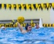 Water polo player prepares to shoot while splashing in a pool, with yellow and black flags in the background.