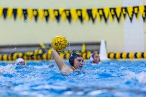 Water polo player prepares to shoot while splashing in a pool, with yellow and black flags in the background.