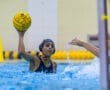 A water polo player throws the ball, splashing water during an intense match in an indoor pool setting.