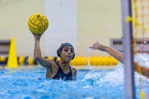A water polo player throws the ball, splashing water during an intense match in an indoor pool setting.