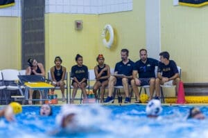 Water polo team during a match, with players and coaches on the sidelines, observing the action in the pool.
