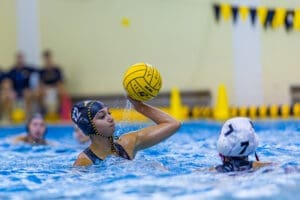 Youth water polo player in action, preparing to throw a yellow ball during a competitive match in a swimming pool.