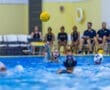 A water polo player prepares to pass the ball during a match, with teammates and coaches in the background.