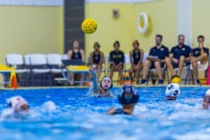A water polo player prepares to pass the ball during a match, with teammates and coaches in the background.