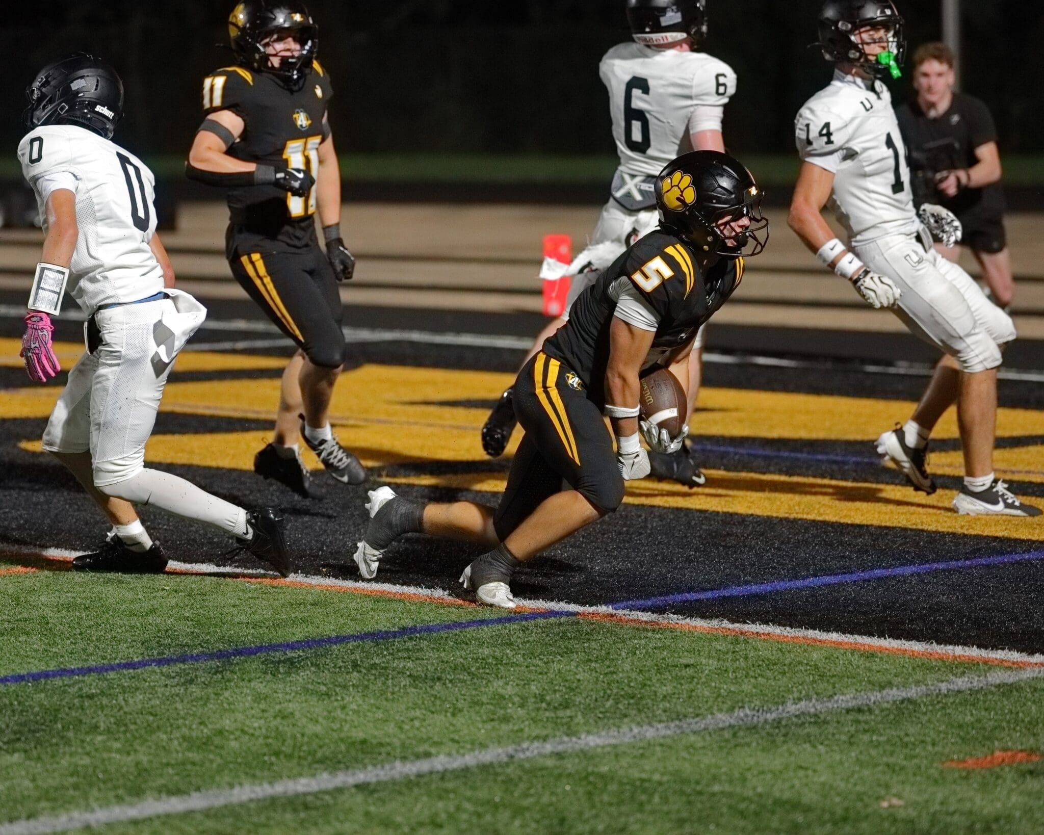 A football player in black uniform runs toward the end zone, evading defenders during a nighttime game.