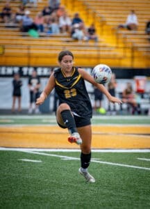 Female soccer player in a black jersey kicking a ball on a grass field, with fans in the background.
