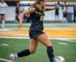 A female soccer player in a black uniform skillfully dribbles a ball on the field during a game.