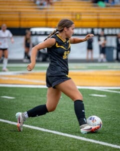 A female soccer player in a black uniform skillfully dribbles a ball on the field during a game.