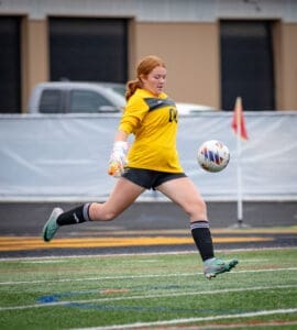 Goalkeeper in action, wearing a yellow jersey, kicking a soccer ball on a field during a game.
