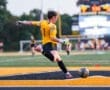 Goalkeeper in action, kicking a soccer ball on a field with a scoreboard in the background during a game.