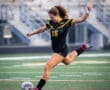 A female soccer player in a black and yellow uniform kicks a soccer ball on a field during a game.