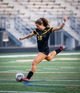 A female soccer player in a black and yellow uniform kicks a soccer ball on a field during a game.