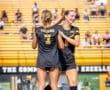 Two female soccer players in black jerseys celebrate together on the field, showcasing team spirit at a game.