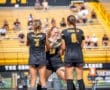 Three female soccer players in black uniforms celebrate on the field during a sunny day at a game.