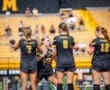Four soccer players in black uniforms, celebrating on the field with a cheering crowd in the background.