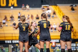 Four soccer players in black uniforms, celebrating on the field with a cheering crowd in the background.