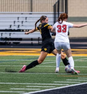 Two high school girls soccer players compete for the ball on a turf field during a match. Intense sports action captured.