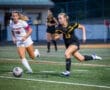 Two high school female soccer players sprinting after a ball on a well-lit field during an evening match.