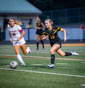 Two high school female soccer players sprinting after a ball on a well-lit field during an evening match.