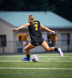 Female soccer player in action, wearing black jersey with Tigers and number 7, kicking a soccer ball on the field.
