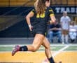 Female soccer player sprinting on a field at night, wearing a black jersey with yellow accents and cleats.