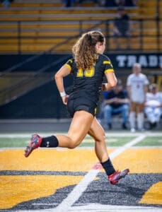 Female soccer player sprinting on a field at night, wearing a black jersey with yellow accents and cleats.