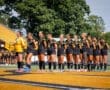 Team lineup of female soccer players in black uniforms, preparing for a match on a sunny day at the field.