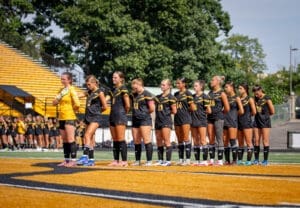 Team lineup of female soccer players in black uniforms, preparing for a match on a sunny day at the field.